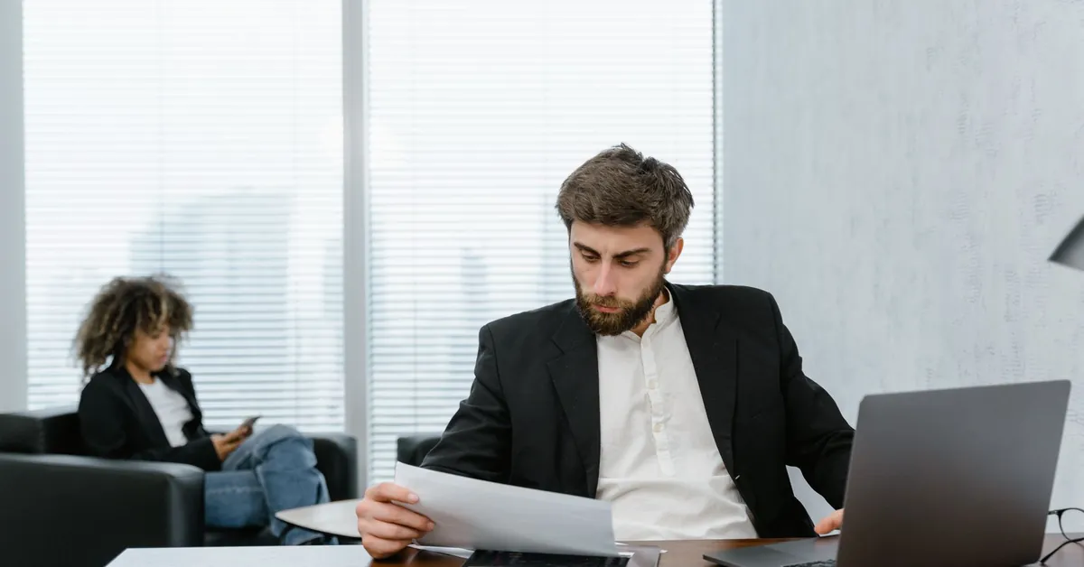 Focused businessman reviews financial reports at office desk with laptop. man in black suit jacket analyzing data reports