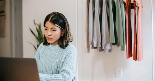 Serious young Hispanic female freelancer in casual outfit surfing on laptop at table near cup with drink at bright home with wardrobe with garments and plant with green leaves concentrated latin american lady working remotely on netbook near wardrobe