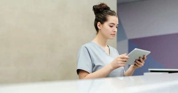 A receptionist in scrubs using a tablet at an office reception desk. a receptionist looking at a tablet