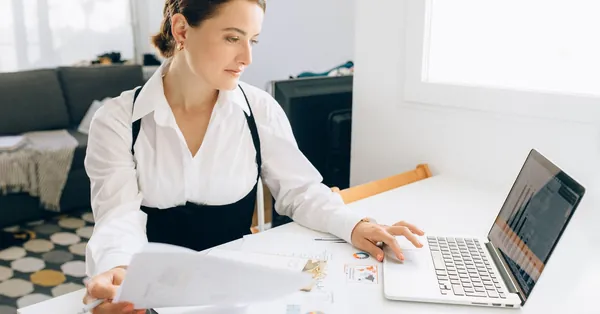 Woman at desk reviewing business reports on laptop, holding papers in a modern office setting. a woman using a laptop