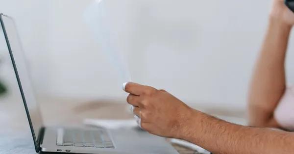 Crop anonymous male reading document and talking on mobile phone while sitting at desk with laptop crop unrecognizable man reading document and talking on phone