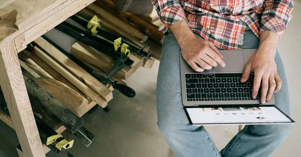 A craftsman using a laptop in a woodworking workshop, focusing on online research or design. person in blue denim jeans sitting on chair using macbook pro