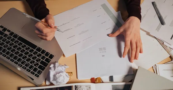 A person taking notes and working on a laptop surrounded by documents on a desk. person holding white document paper