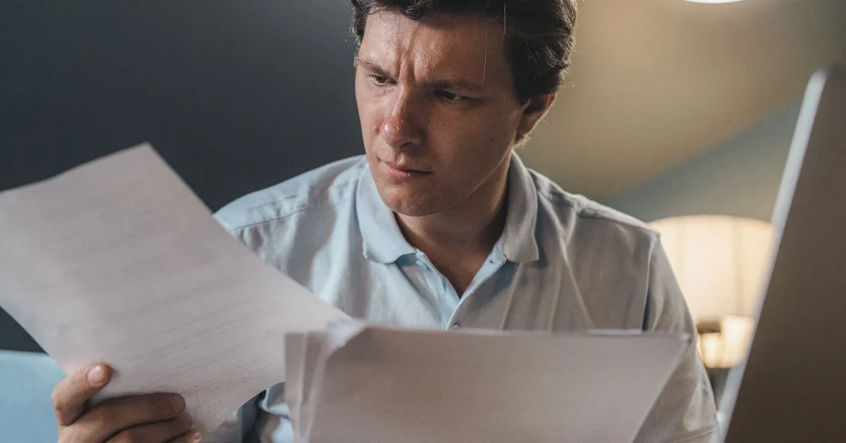 A man intently examines papers, seated indoors under warm lighting, focusing on his work. a man holding printer papers