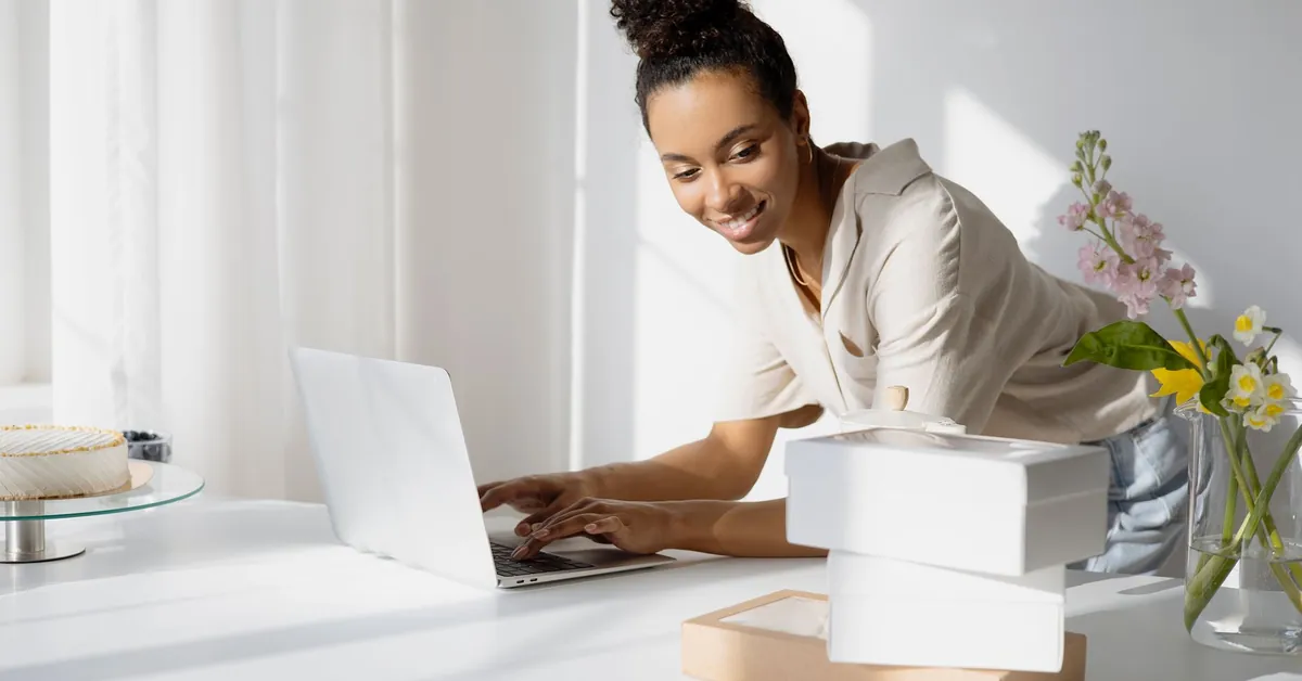 African American woman working on a laptop with boxes and flowers in a bright room. a woman typing while looking at boxes