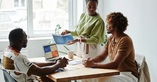 Three colleagues engage in a collaborative meeting, discussing a project in a bright, modern office. woman holding a paper while talking to her colleagues